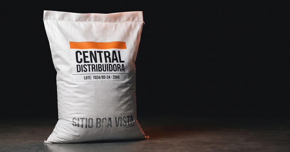 Saca de ráfia branca sobre concreto polido escuro, com um carimbo pequeno e desbotado do produtor perto da barra, escrito SITIO BOA VISTA, e uma etiqueta industrial grande e nova colada por cima da frente, escrita CENTRAL DISTRIBUIDORA, com tarja laranja e código de lote